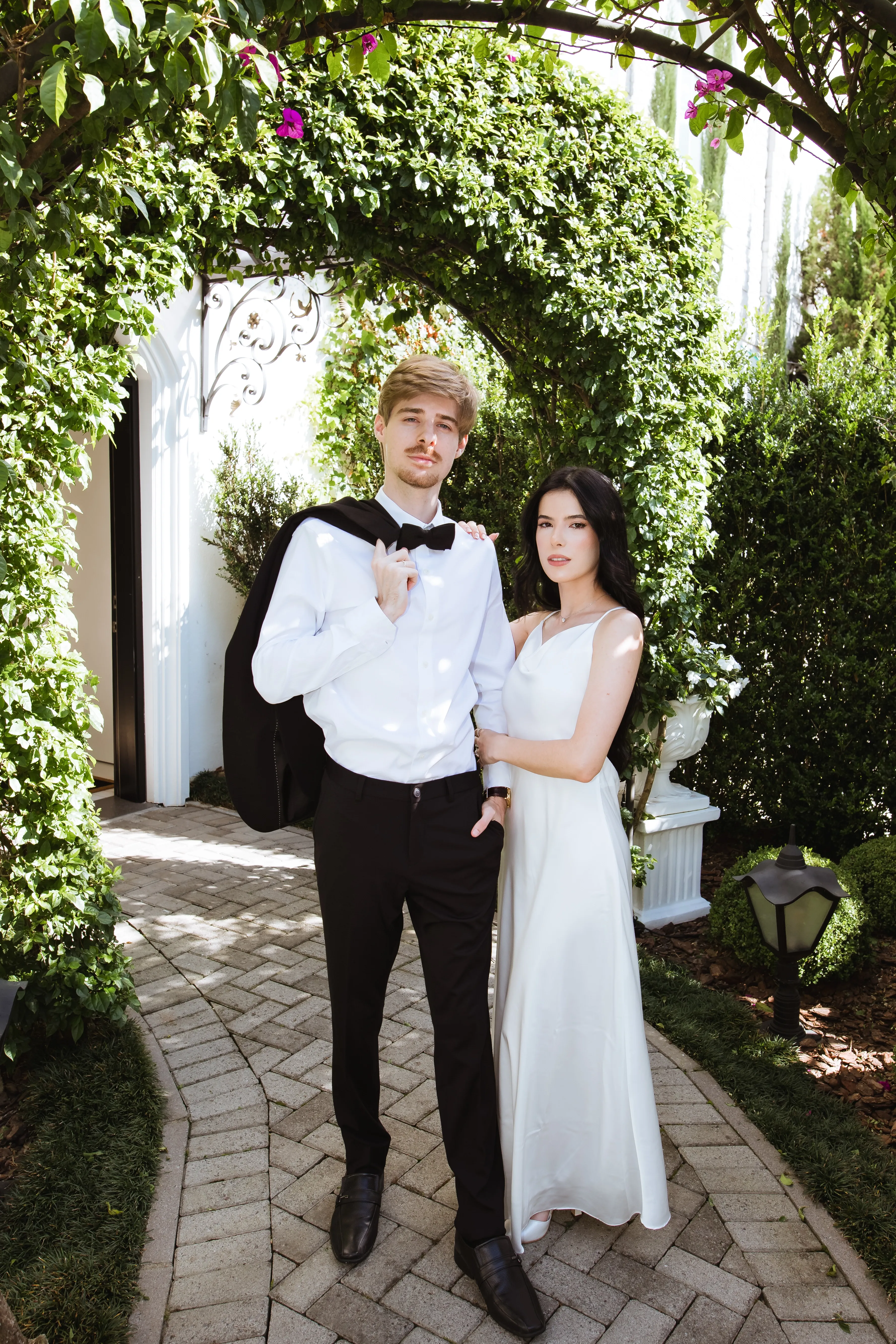 Rafael e Liliane posando com trajes elegantes em um jardim parcialmente iluminado pelo sol. Rafael está à esquerda, vestindo camisa branca, calça preta e sapatos pretos, com seu terno preto jogado sobre o ombro. Lili está à direita, olhando diretamente para a câmera em um belo vestido longo branco, apoiando uma mão no ombro de Rafael enquanto a outra envolve seu braço.